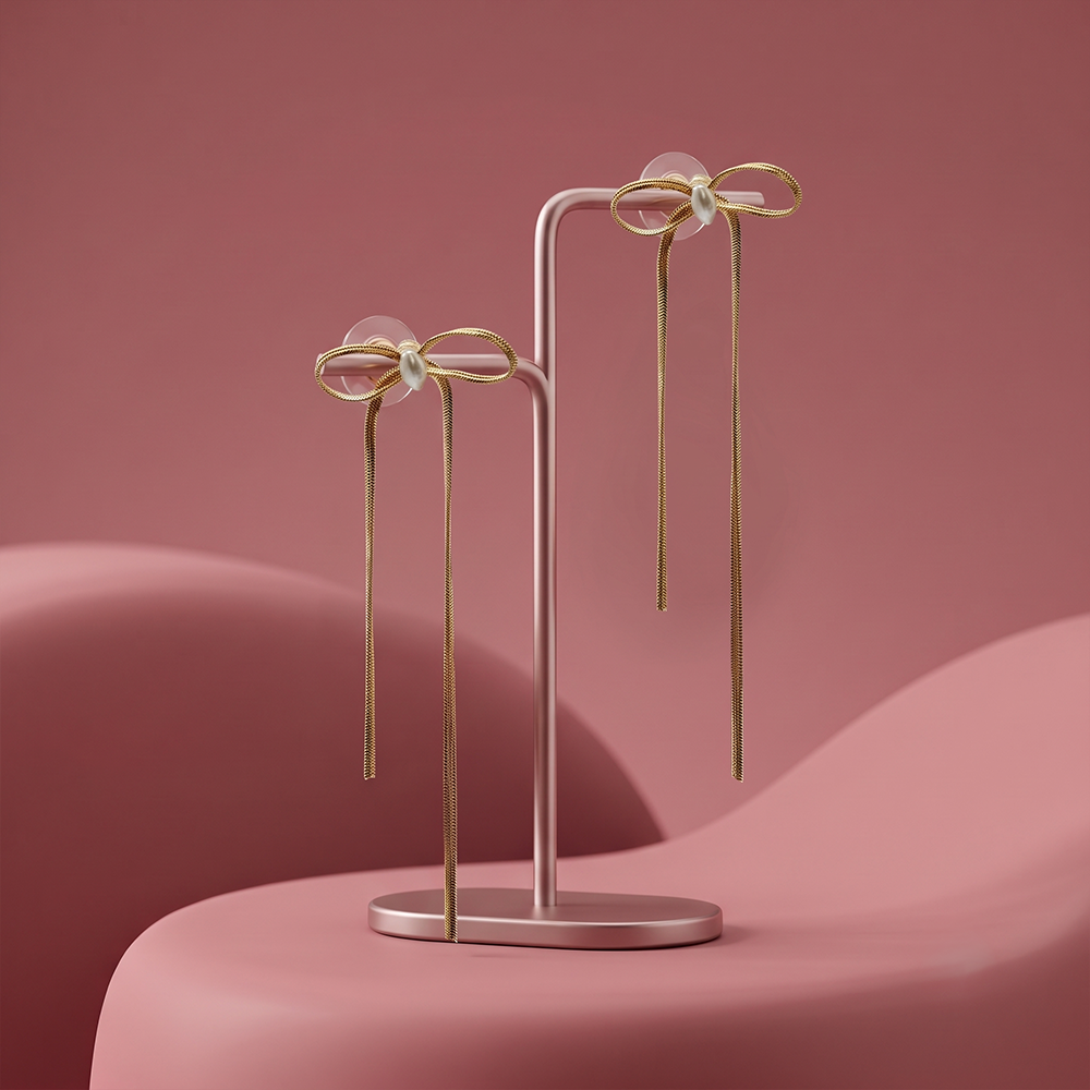 A pair of gold bow dangle earrings with central pearls displayed on a minimalist pink jewelry stand against a pink background.