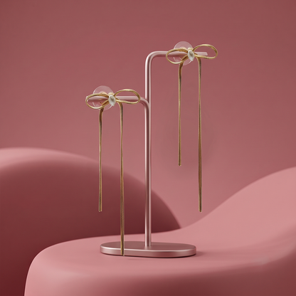 A pair of gold bow dangle earrings with central pearls displayed on a minimalist pink jewelry stand against a pink background.